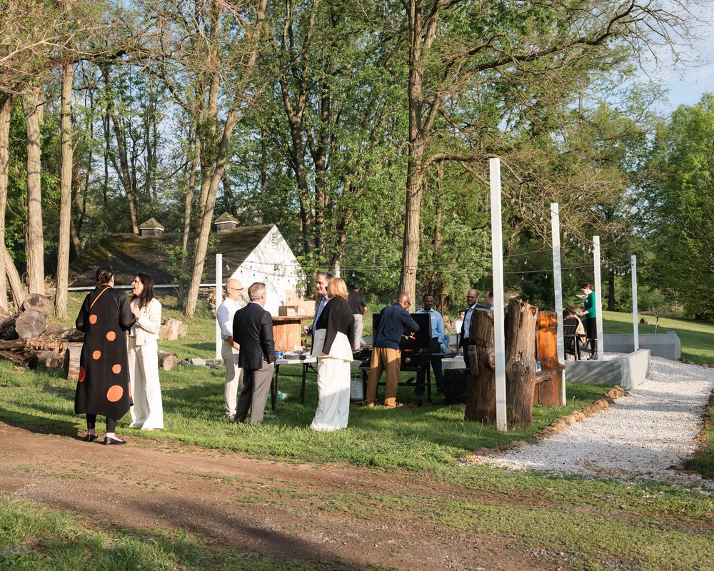 Guests mingling at an outdoor event near wooden sculptures and tall trees in a scenic park setting.