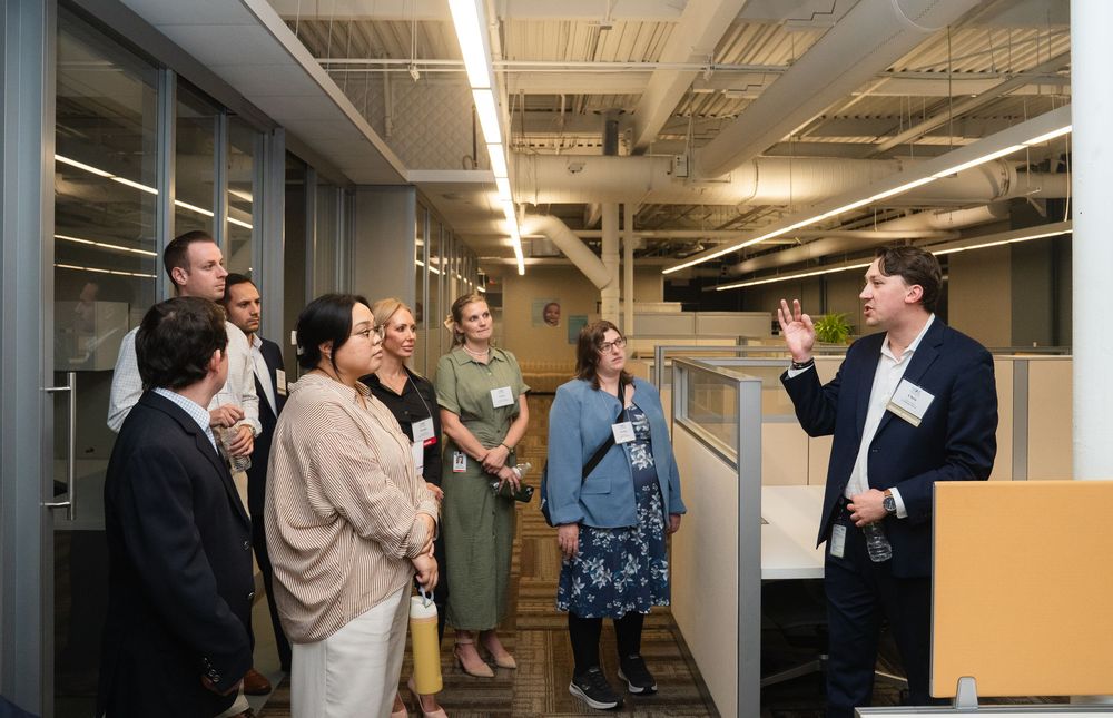People interact and network in a modern office hallway with glass walls and fluorescent lighting.