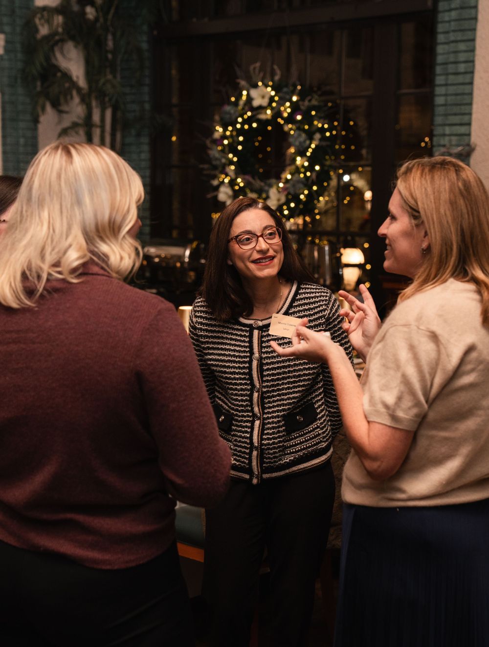 Three women socializing at a festive holiday party with a decorated Christmas tree glowing in the background.