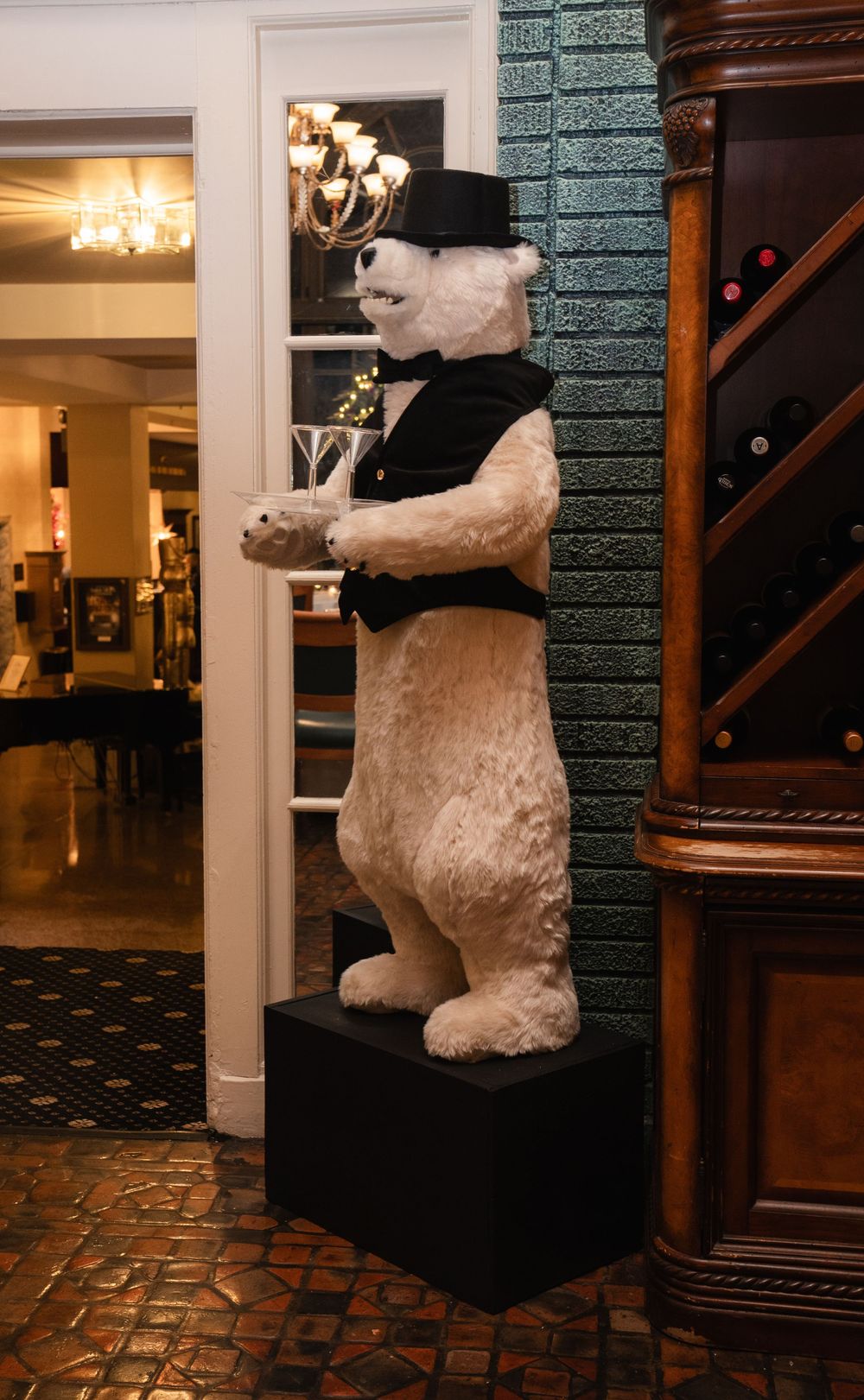 Large polar bear mascot statue wearing a top hat and vest, holding a tray, standing on a pedestal in an elegant restaurant.