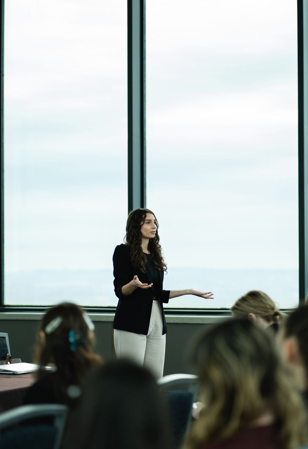 A presenter speaks to an audience in a modern conference room with large windows and natural lighting.