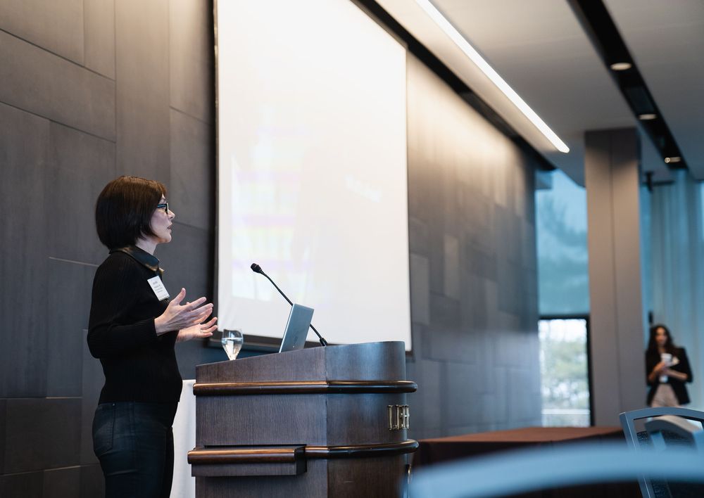 A speaker presents from a podium at a professional conference or event in a modern venue.