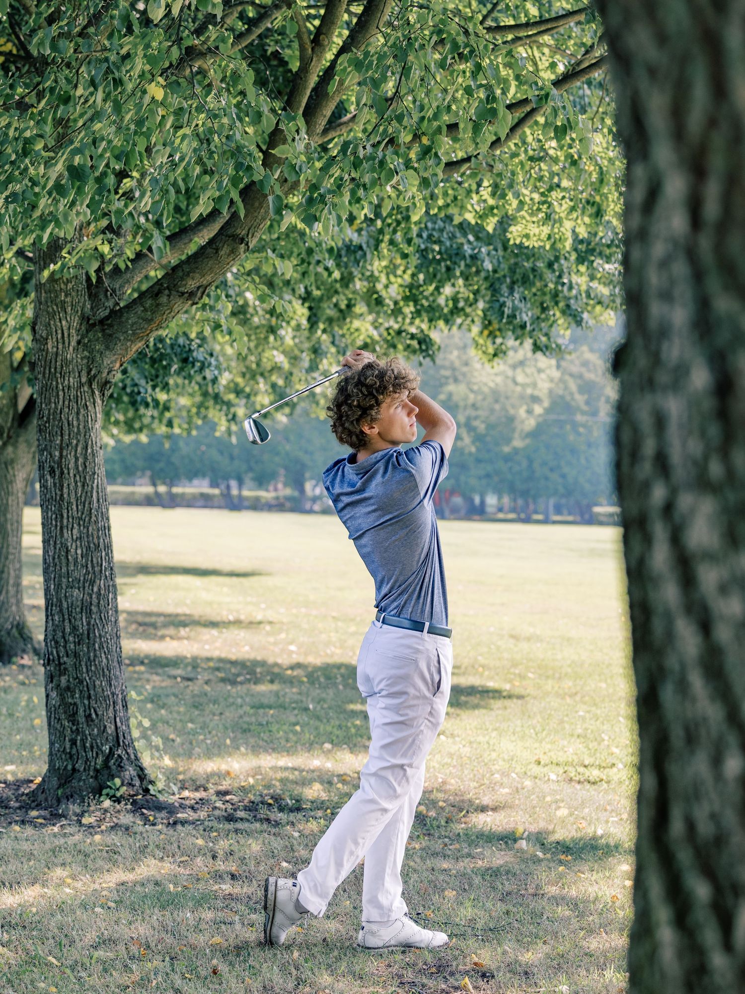 Golfer in blue shirt and white pants takes a full swing on a sunlit course framed by mature trees.