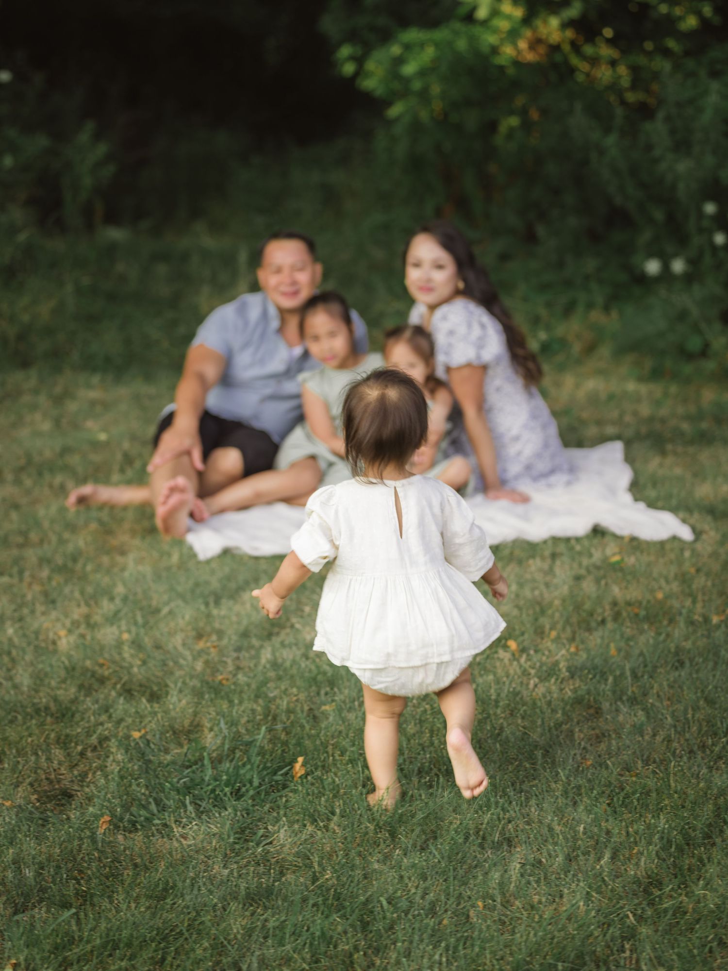 A little one in white clothing walking on grass towards others seated on a blanket for a summer outdoor photoshoot.
