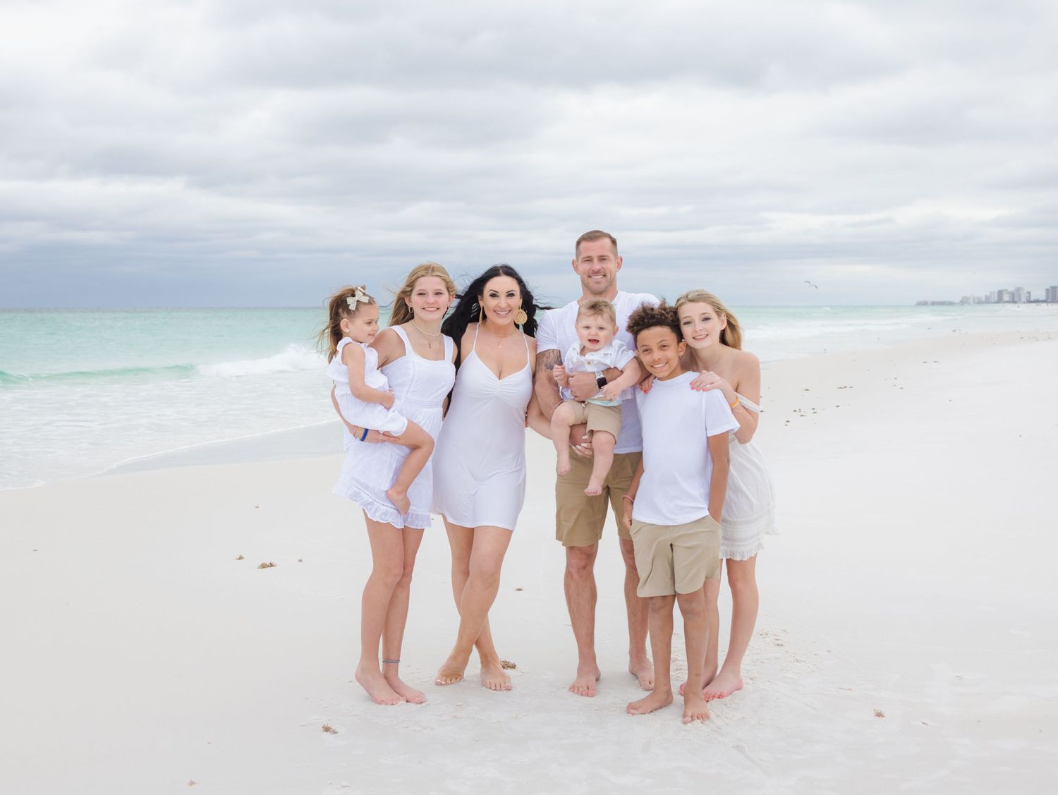 A family in white outfits stands together on a pristine beach with turquoise water and cloudy skies in the background.