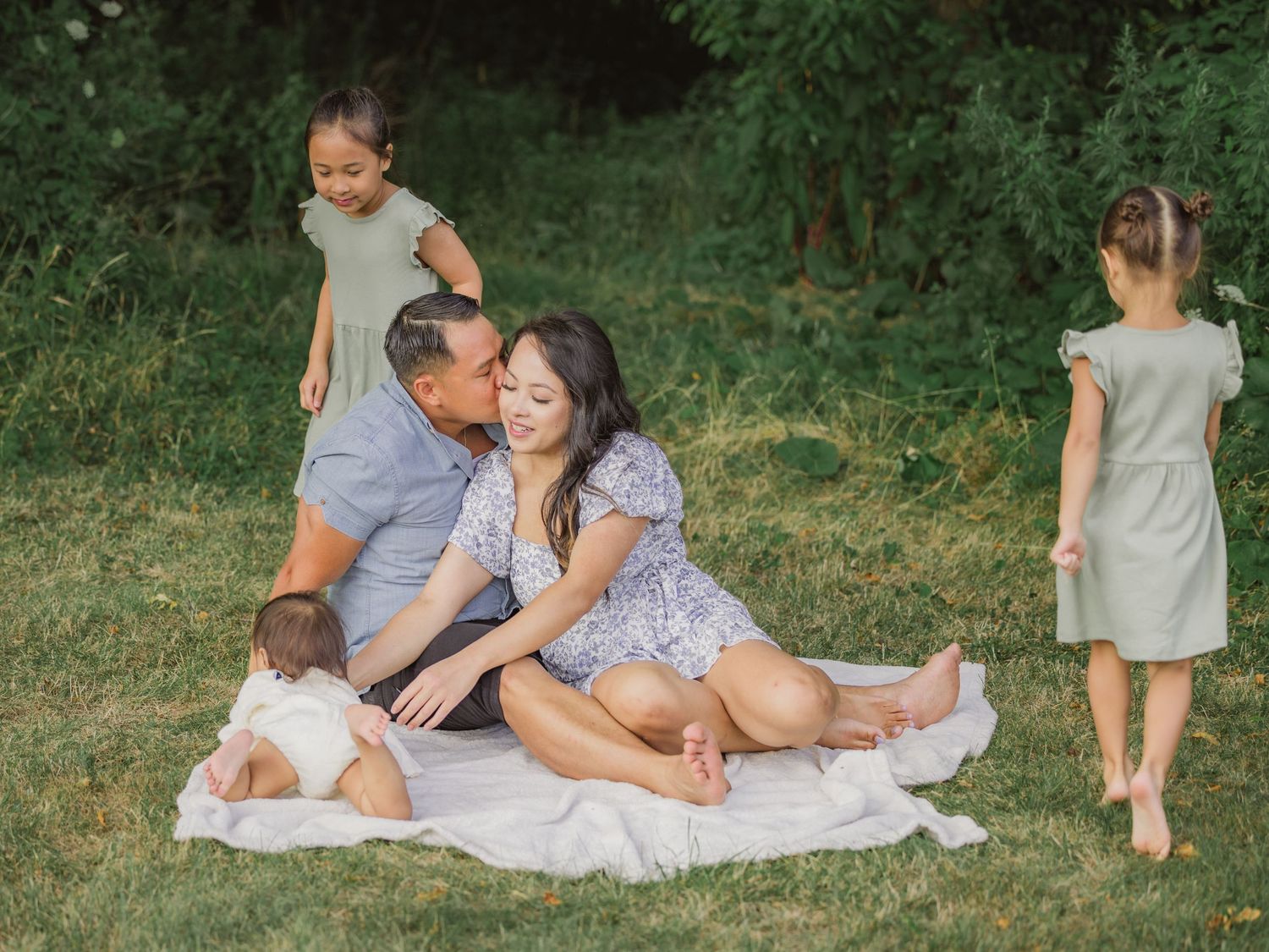 Family enjoying a summer picnic outdoors on a white blanket with green grass and trees in the background.