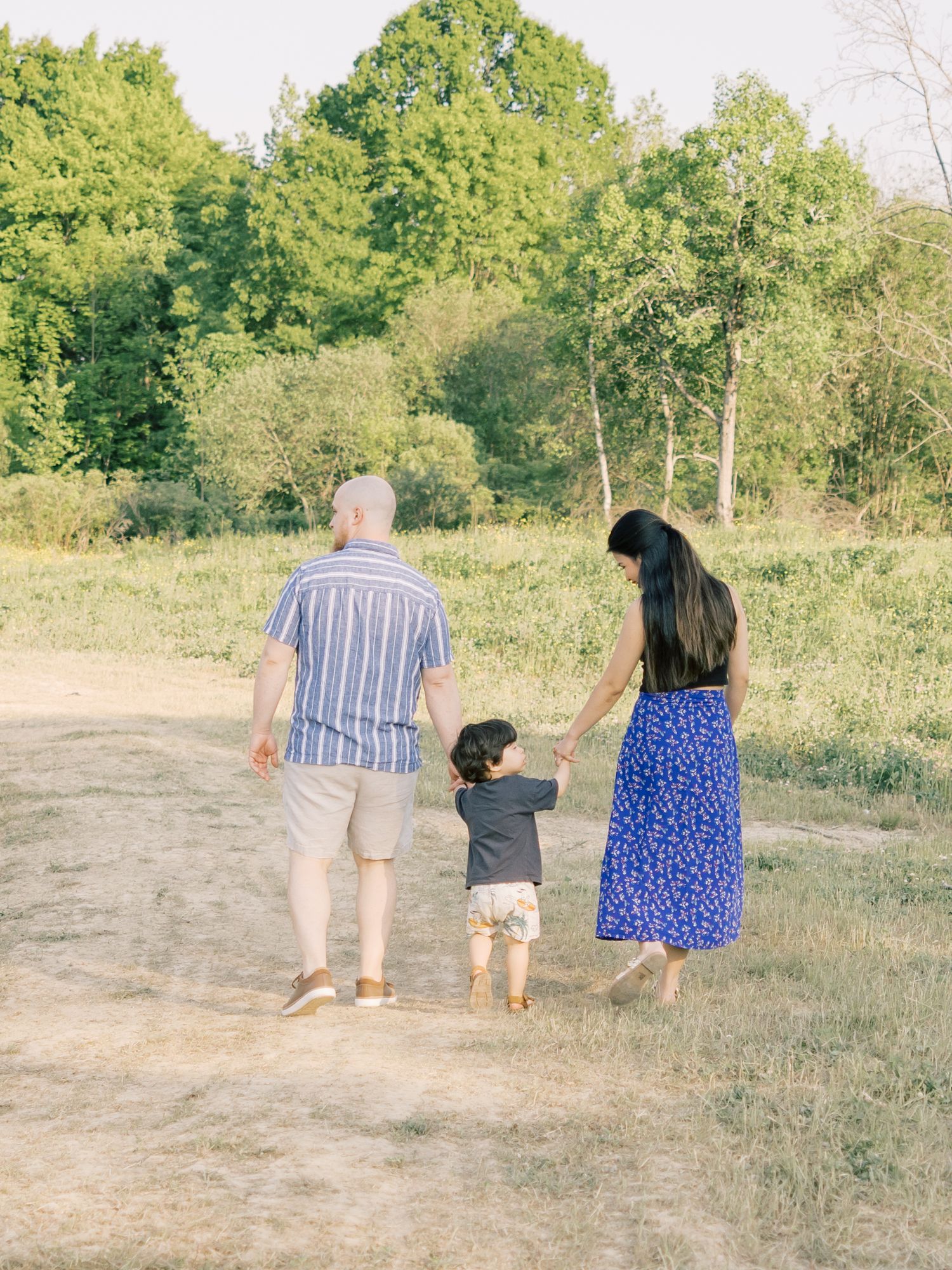 A family takes an outdoor stroll together on a dirt path surrounded by lush green trees on a sunny day.