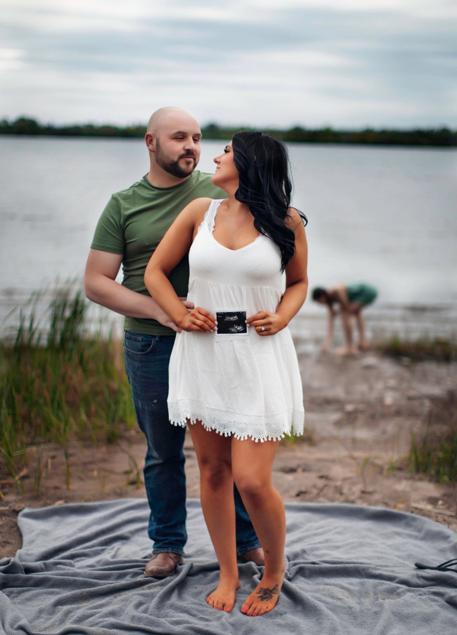 A couple shares an intimate moment on the beach by a lake while holding a sonogram picture.