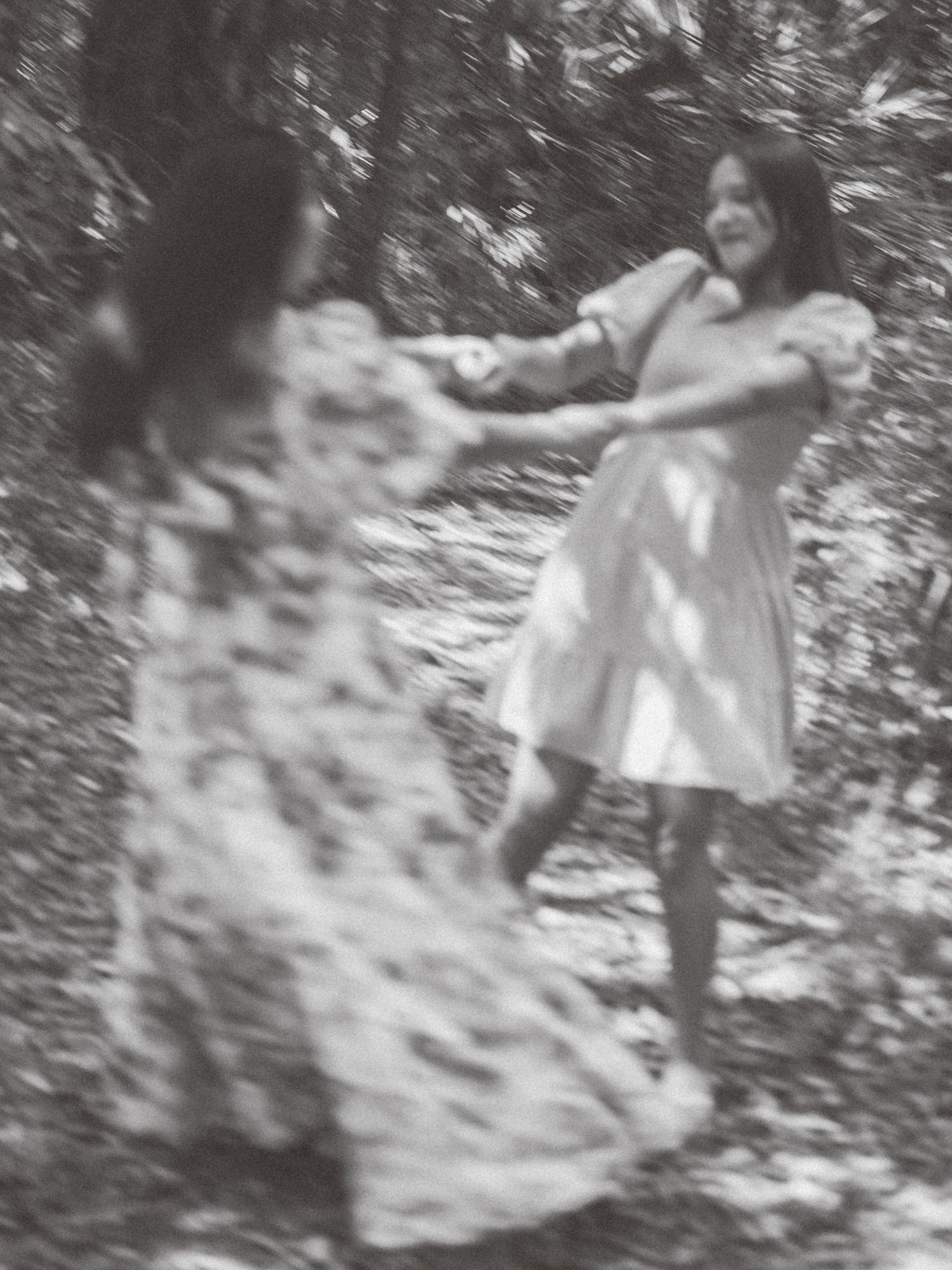 Blurred black and white vintage photograph showing someone in a light dress dancing or spinning in a wooded area.