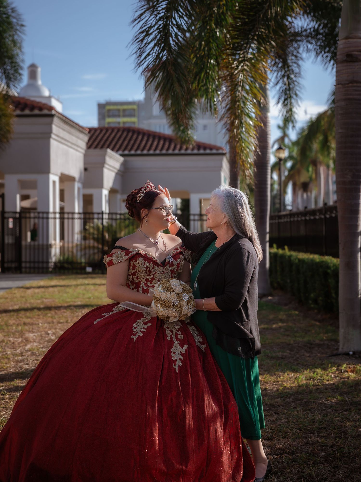A touching moment outdoors with a red quinceañera ballgown and evening wear against palm trees and Spanish-style architecture.
