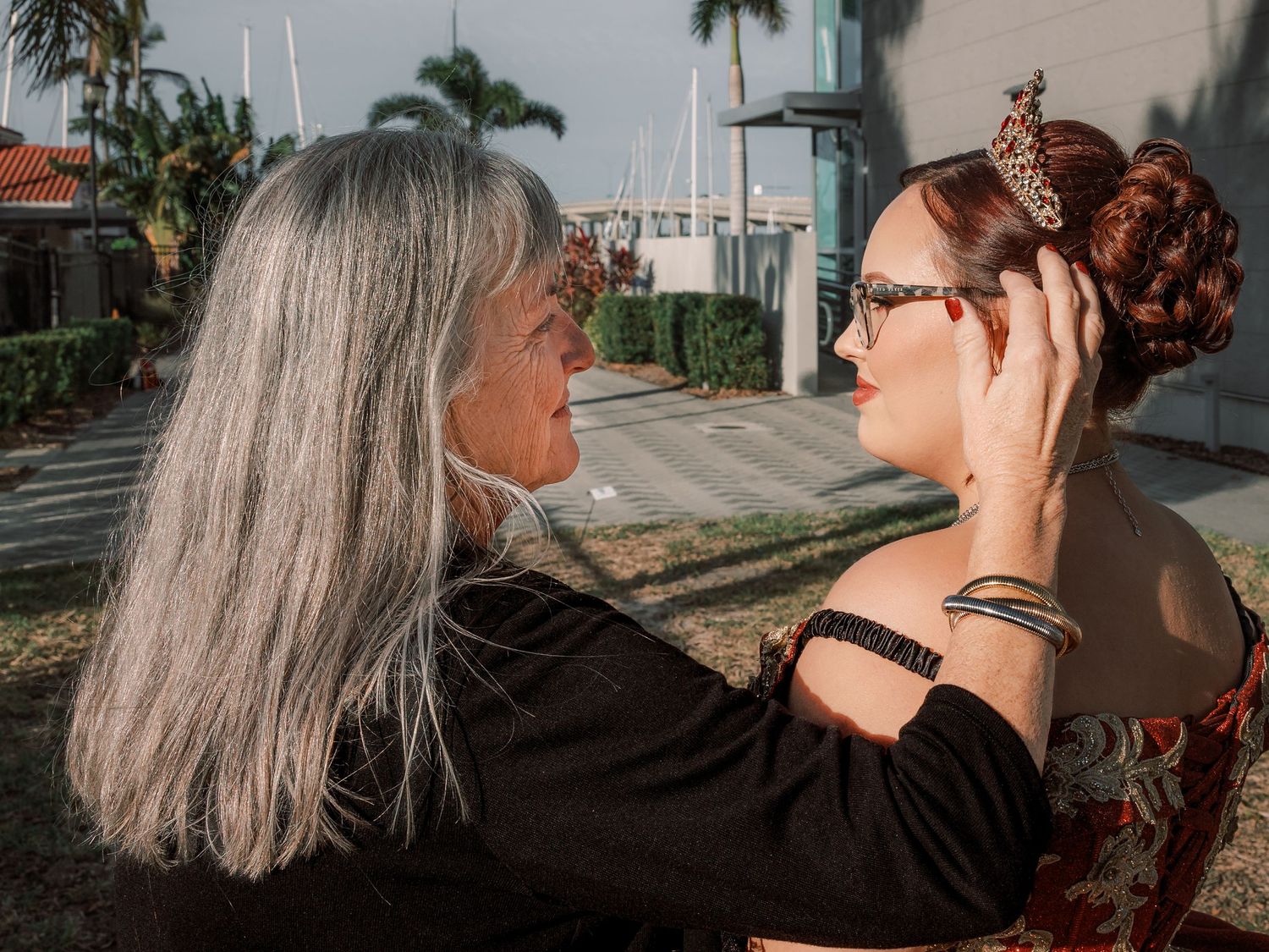 Two women stand together on a palm-lined street, one with long grey hair gently touching the other's styled updo.
