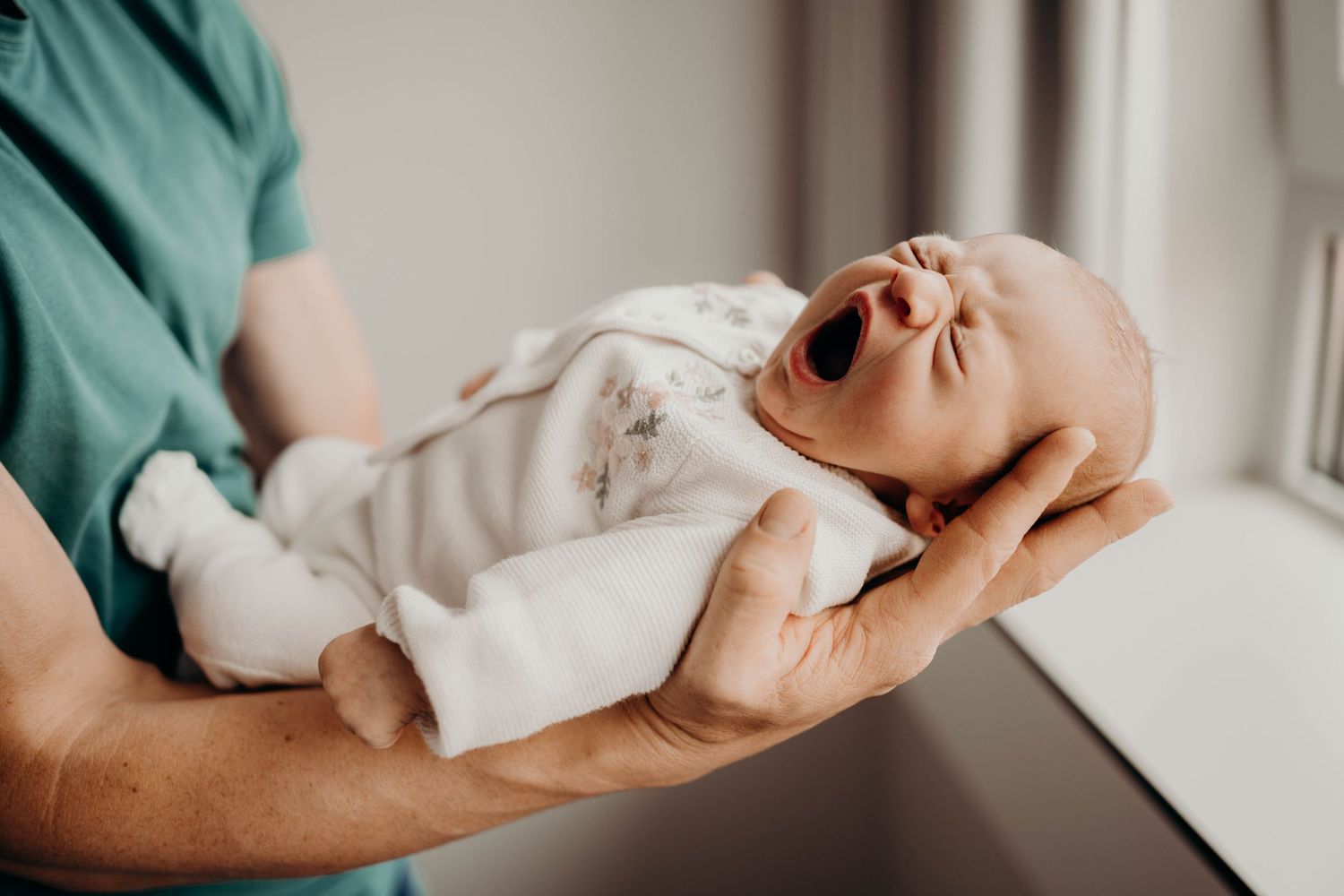 A crying newborn baby being held in gentle hands while wearing a light colored onesie.