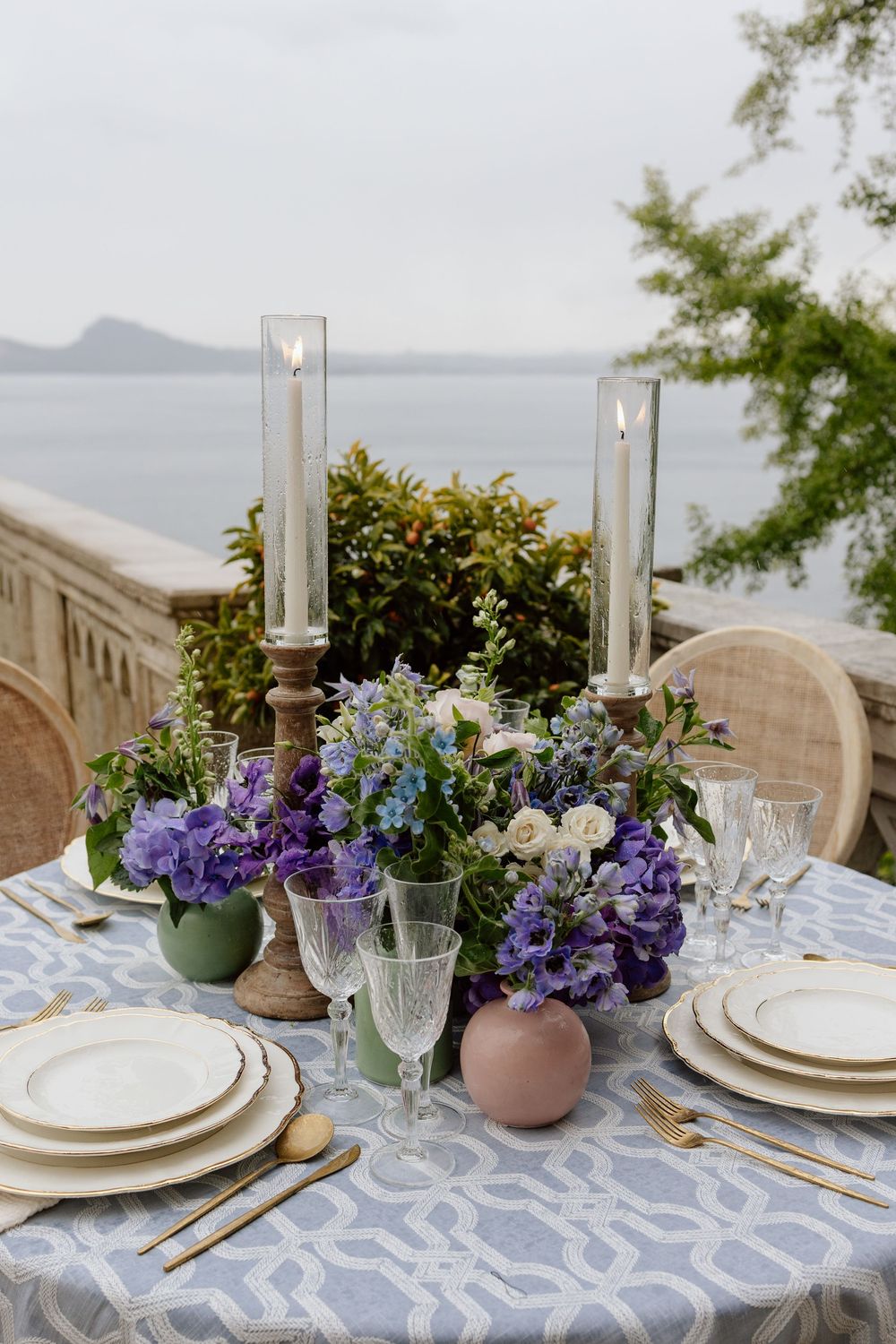 Elegant outdoor table setting with purple hydrangeas, glass candlesticks, and white plates overlooking a tranquil lake view.