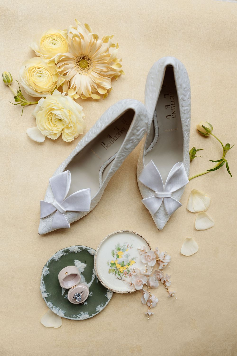 Elegant white bridal flats with bow details arranged with pale yellow ranunculus flowers and vintage decor plates on beige background.
