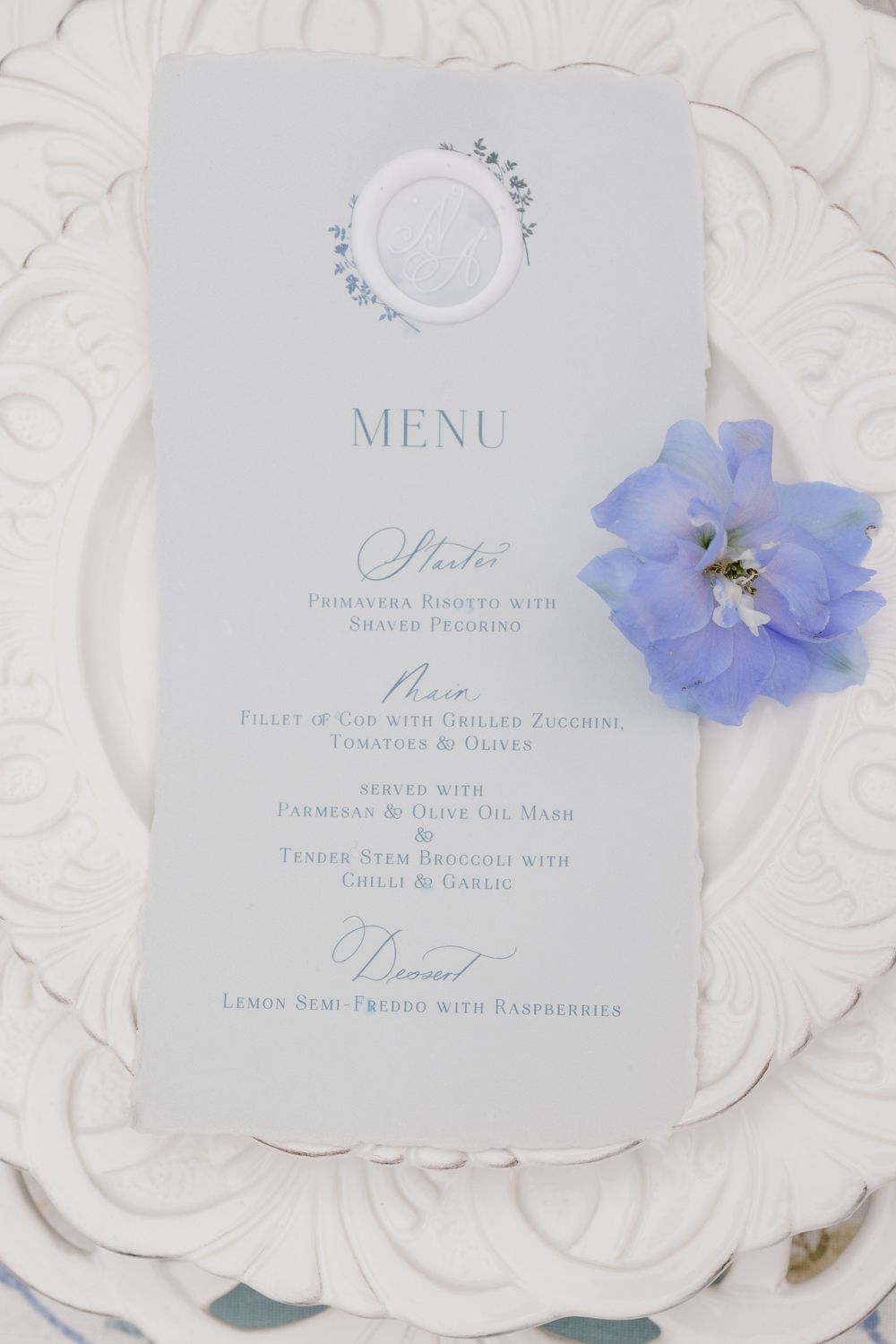 A delicate white menu card with blue floral accents and elegant calligraphy styling features a pale blue delphinium flower.