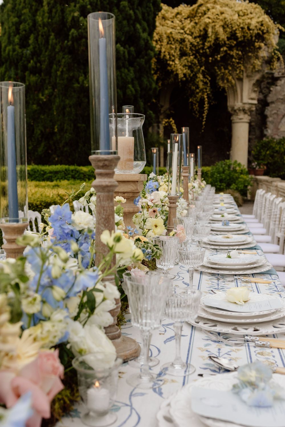 Elegant outdoor wedding reception table with tall candle holders, delicate blue and white floral arrangements, and crystal glassware.