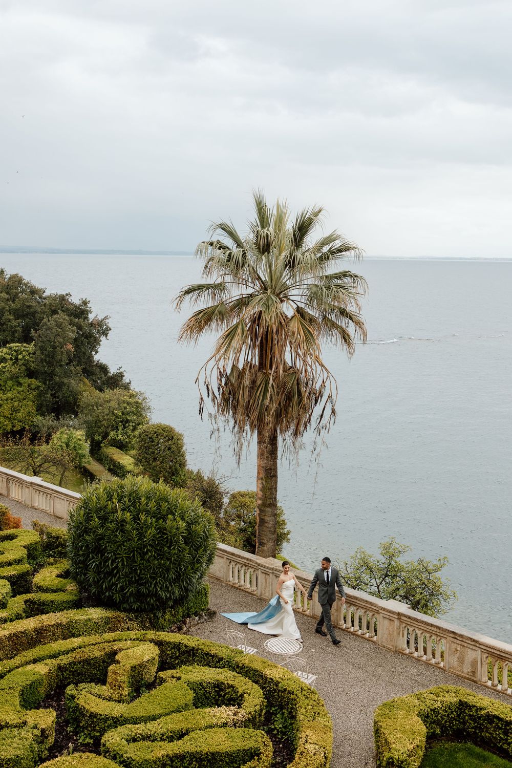 Ornate garden with palm trees and geometric hedges overlooking the ocean from a stone terrace balustrade.