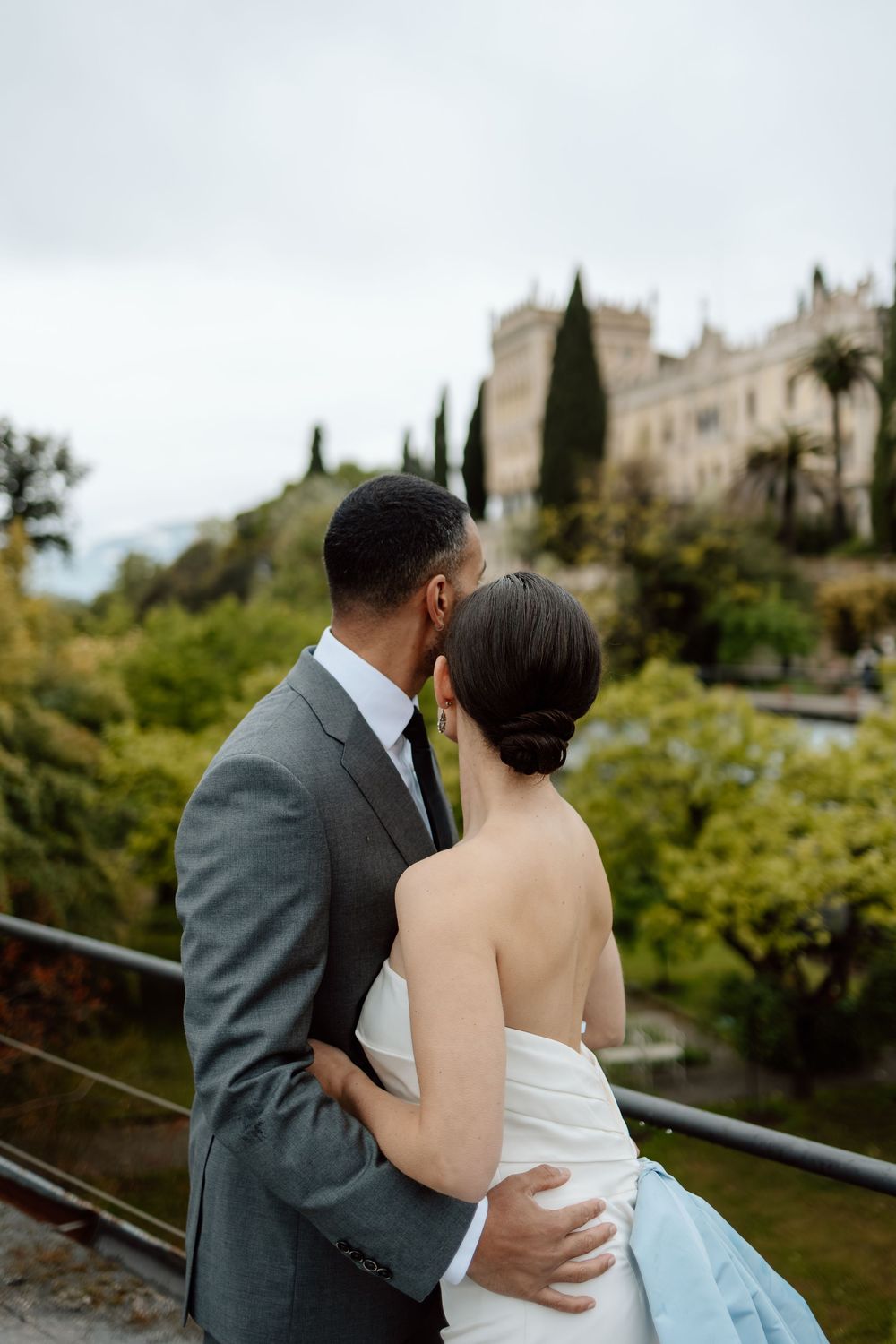 A couple in wedding attire share an intimate moment together on a scenic overlook with lush greenery and buildings behind them.