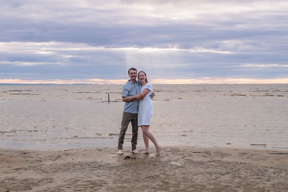 Couple souriant collé l’un à l’autre à la plage de Saint-Gédéon, rayons de soleil perçant les nuages.