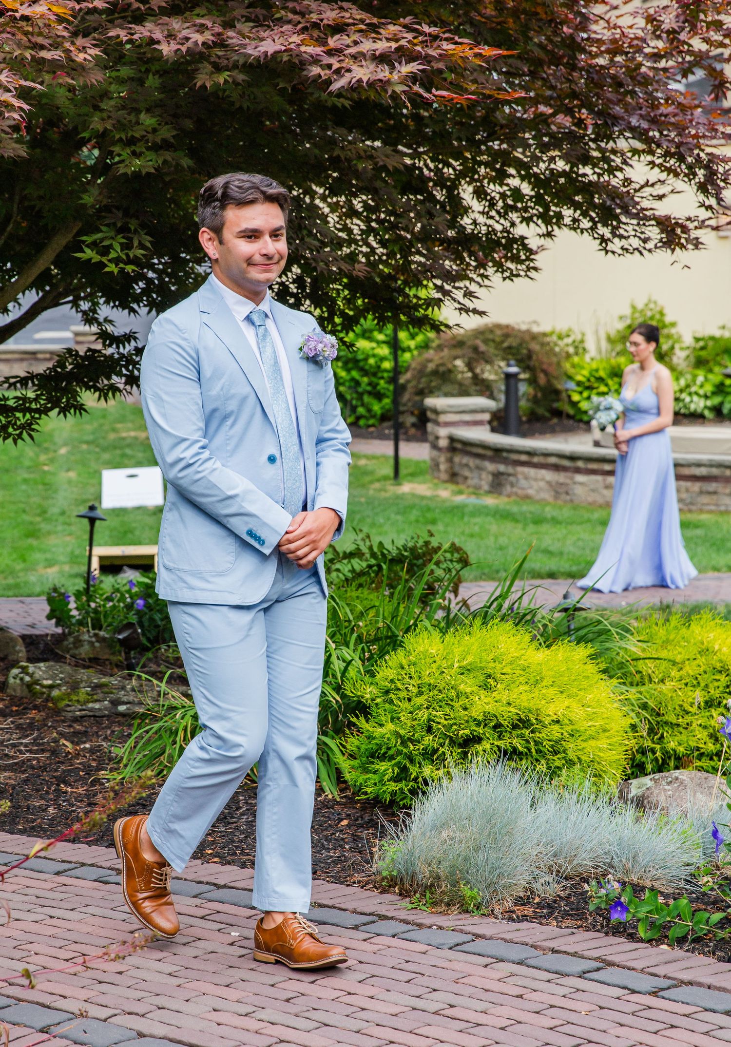 Wedding party photo in a garden setting with light blue suit and long dress against lush greenery and brick pathway backdrop.