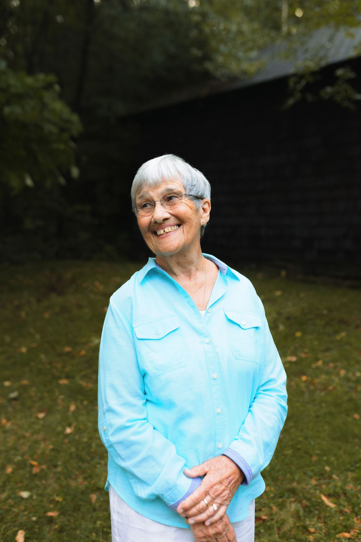 An elderly person wearing a light blue button-up shirt sits outdoors on grass while smiling warmly at the camera.