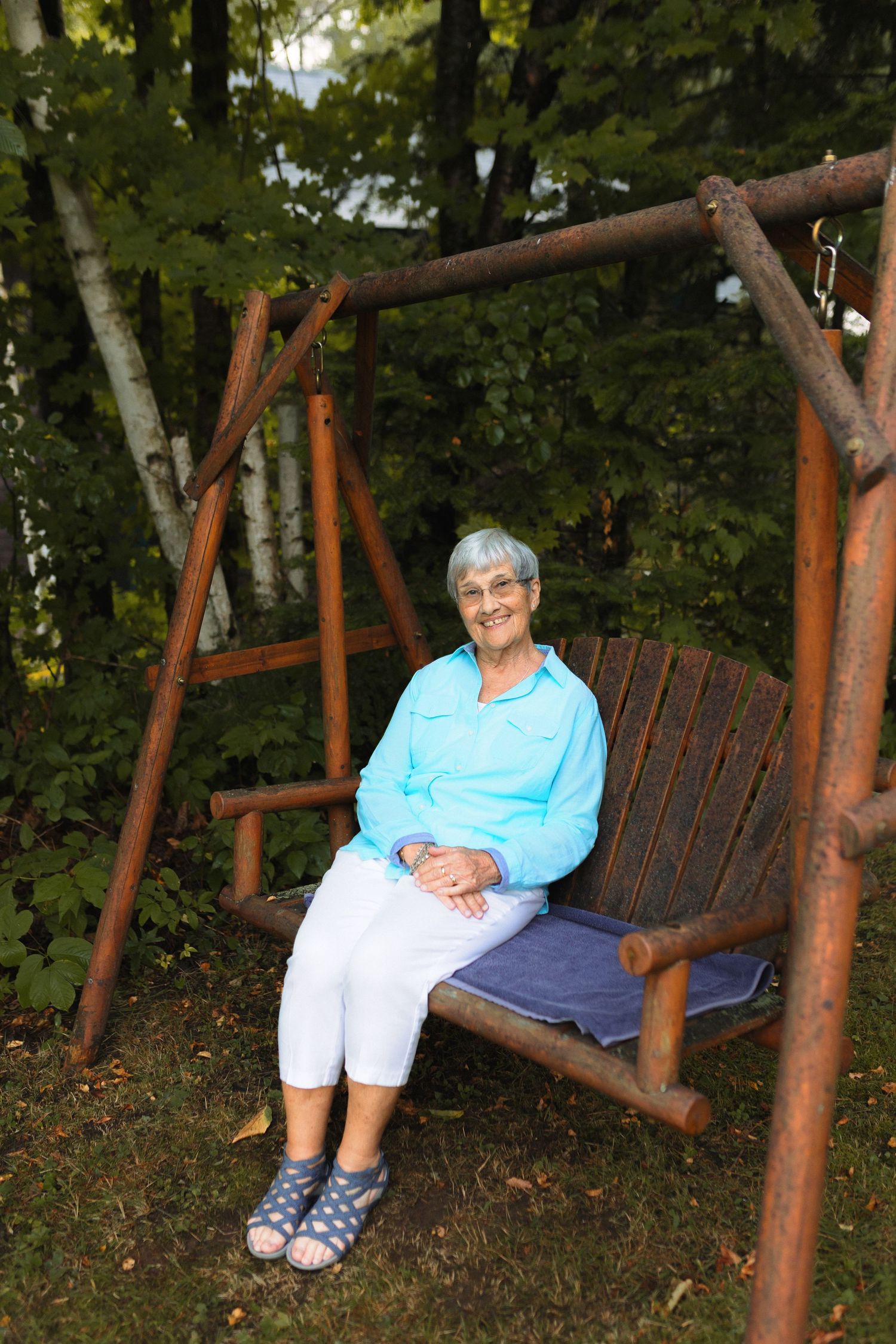 A wooden porch swing hangs in a peaceful wooded setting on a summer day.