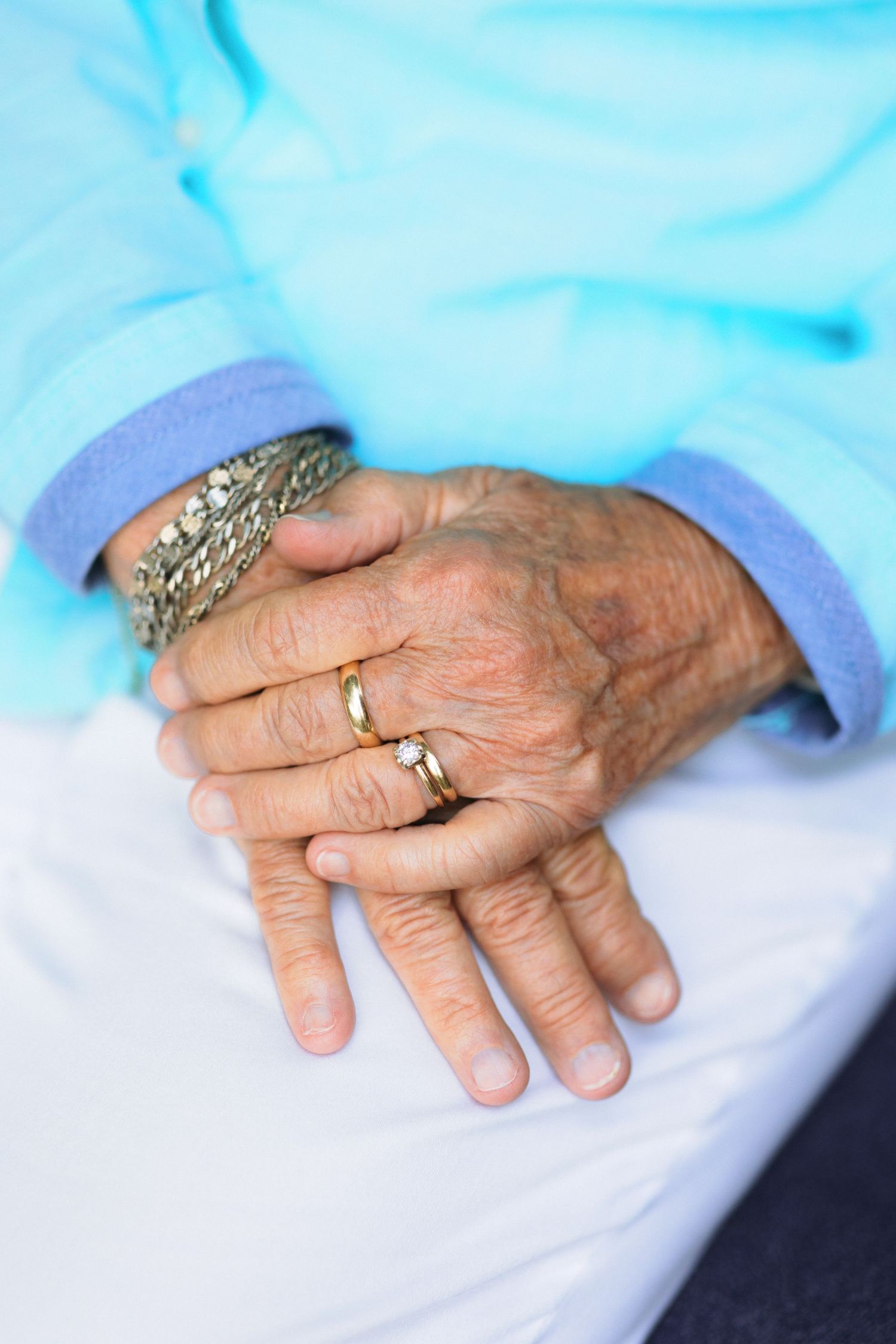 Close-up view of an elderly hand wearing a wedding ring and decorative jewelry against a light blue and white background.