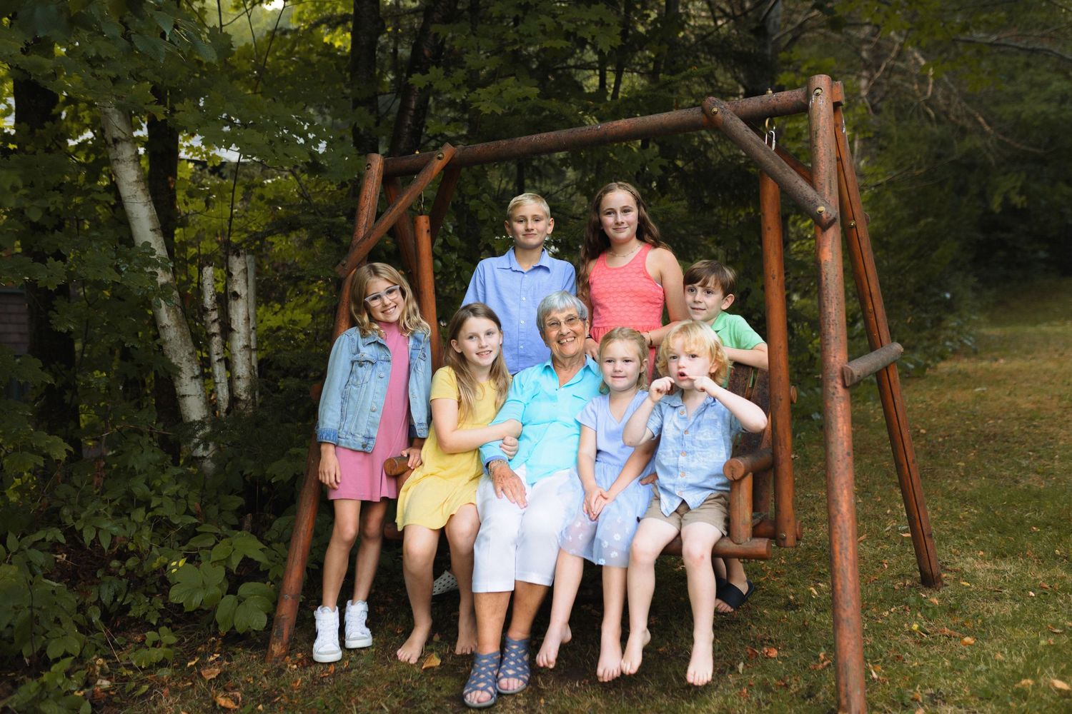 A wooden swing set stands in a green backyard surrounded by trees during a summer day.