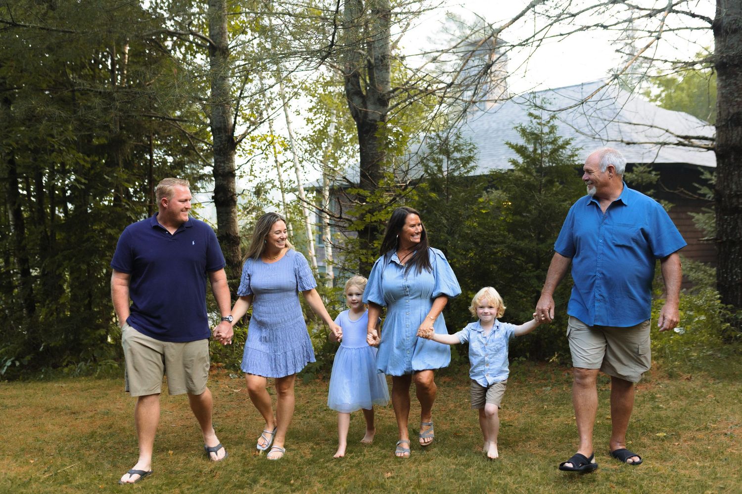 A group in coordinating blue outfits holds hands while standing together on a grassy hill with trees in the background.
