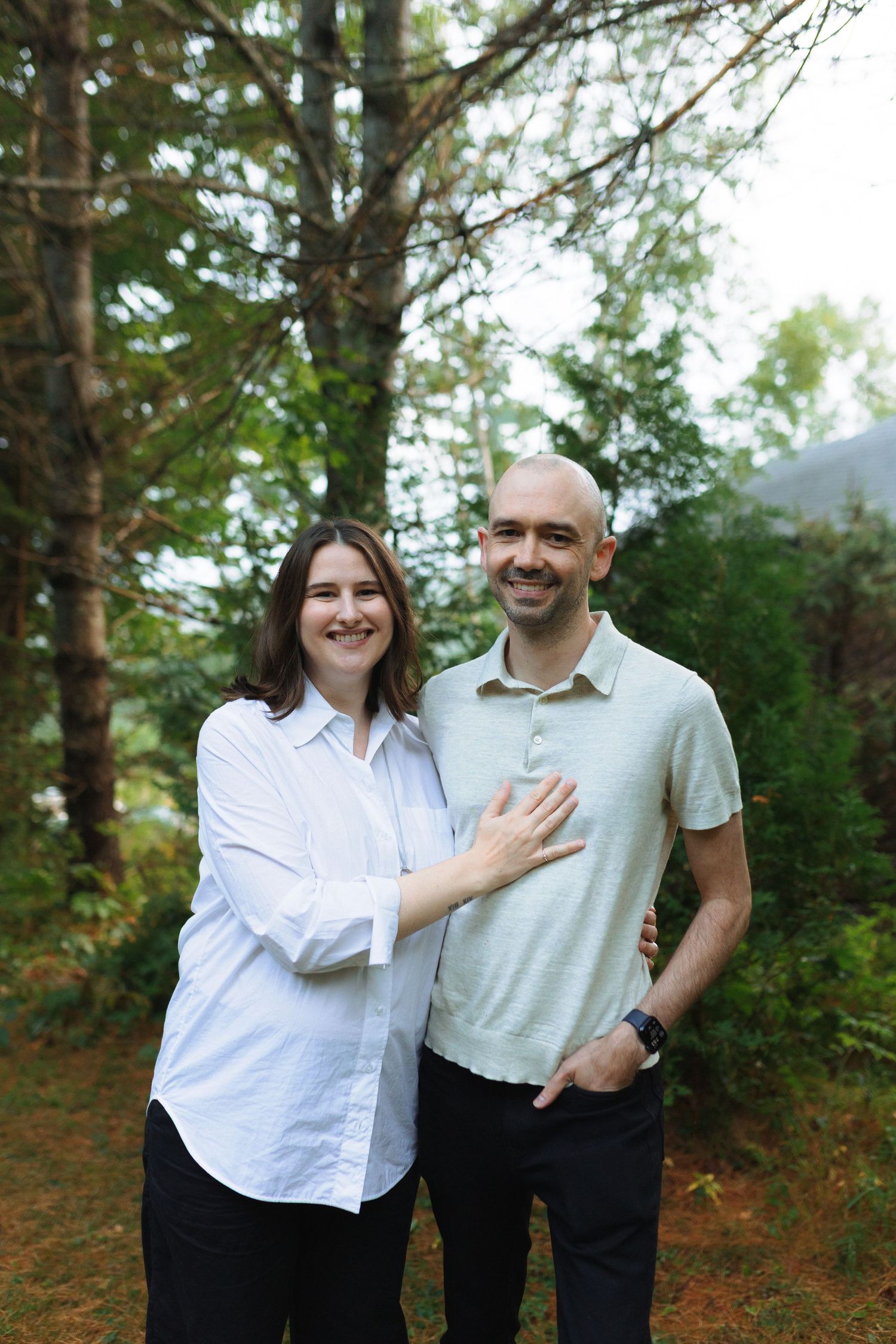 A couple shares joyful laughter while posing together outdoors among tall evergreen trees.