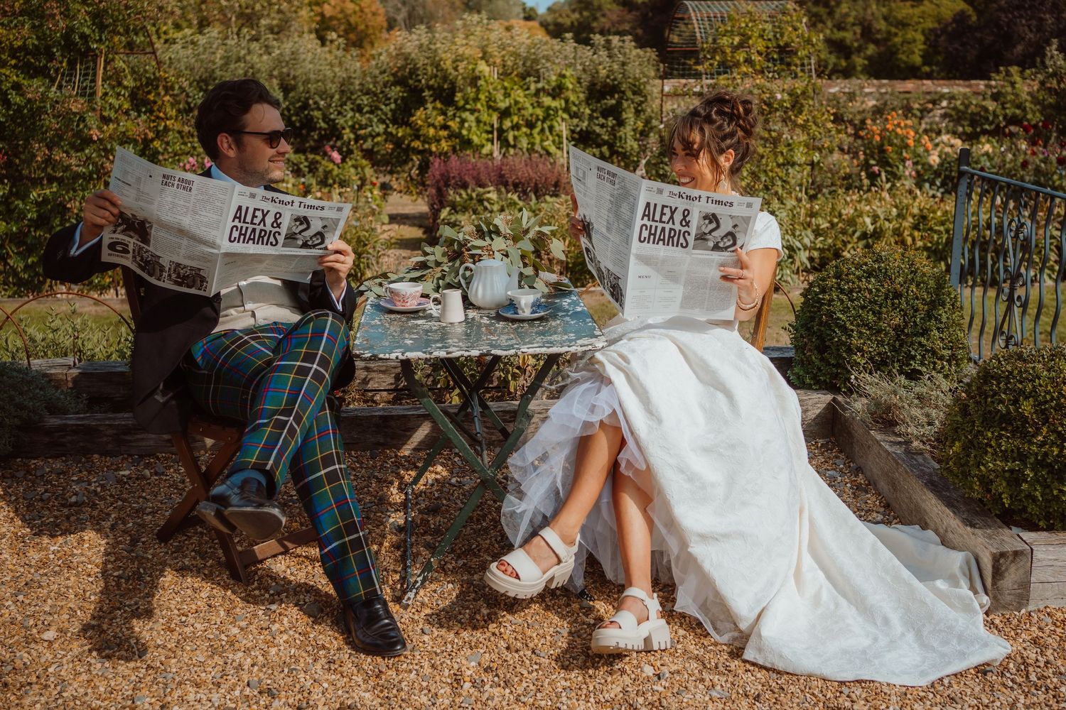 A couple in wedding attire reads newspapers while enjoying an outdoor tea service in a sunny garden setting.