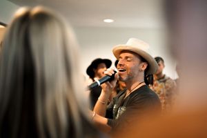 Performer in a hat speaking into microphone during indoor event.