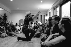 Performer kneels dramatically in front of seated audience members during indoor show.