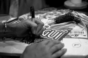 Black and white closeup of hands writing or drawing on paper near corn cob.