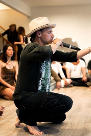 Workshop instructor in hat crouches while speaking into microphone during dance class.