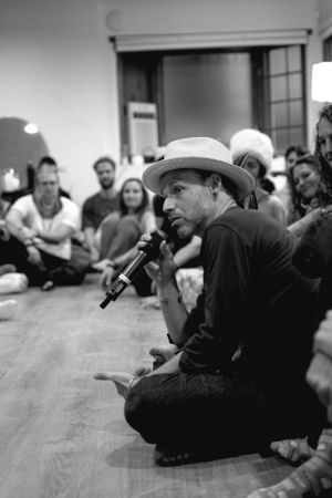 A performer wearing a cowboy hat speaks into a microphone while sitting cross-legged on a floor during an intimate gathering.