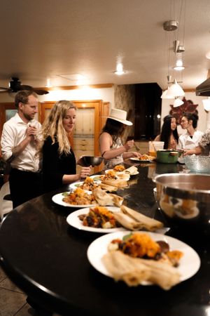 People gather around a table filled with plates of food in a warmly lit indoor setting.