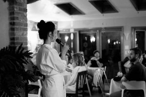 A person in white clothing addresses a seated audience in an indoor venue with exposed brick walls.