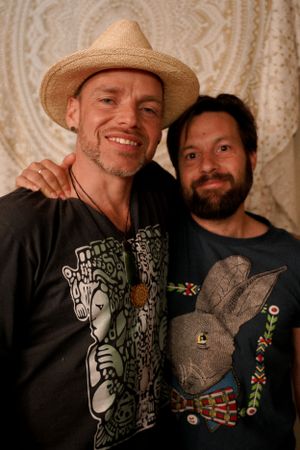 Two friends pose together against a patterned backdrop, one wearing a straw hat and graphic t-shirt.
