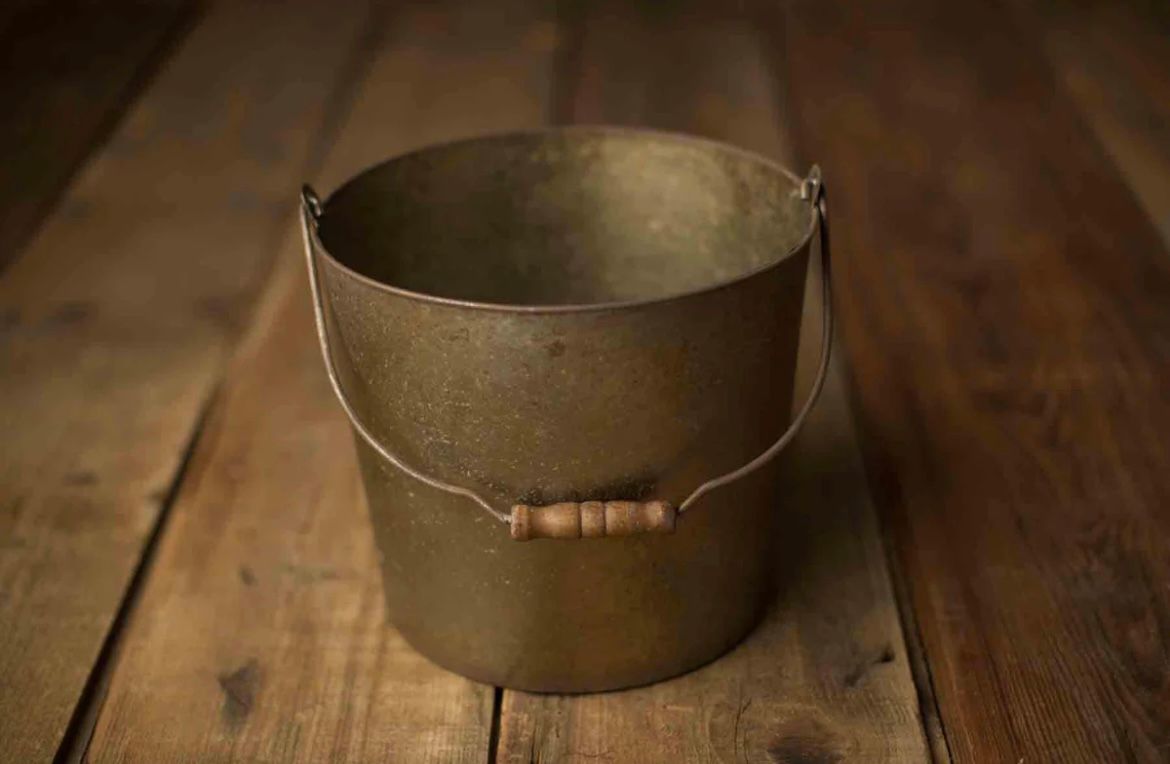Vintage metal bucket with wooden handle sits empty on rustic wooden floorboards.