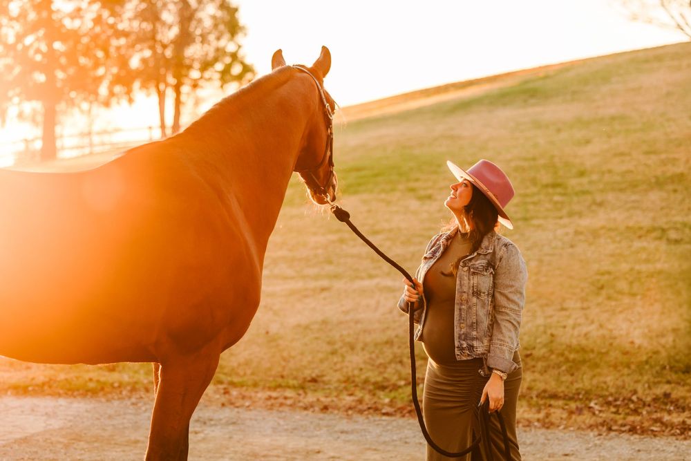 A chestnut horse stands beside a person in a cowboy hat during golden sunset on a grassy hillside.