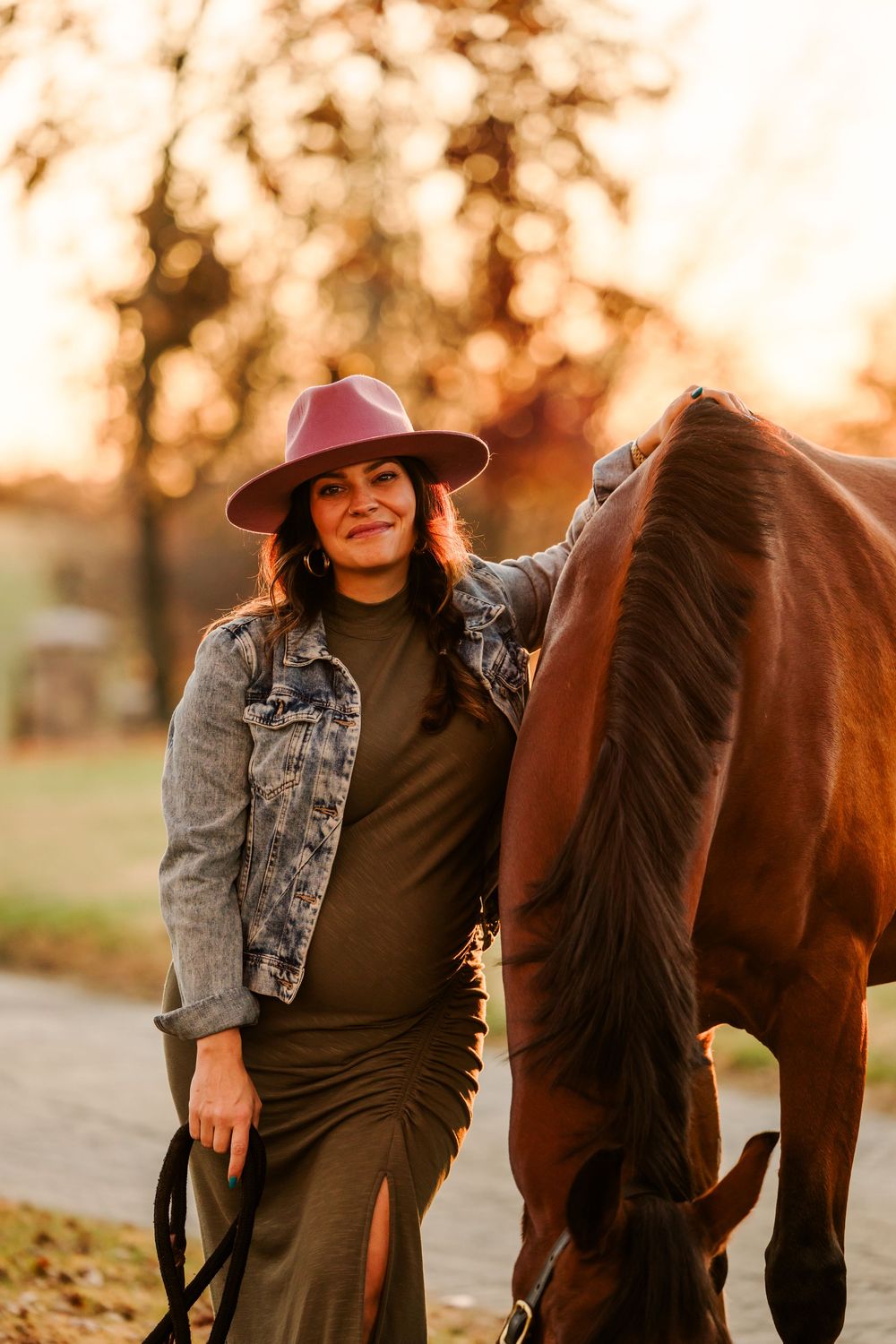 A brown horse grazes while someone wearing a burgundy hat and denim jacket stands beside it at sunset.
