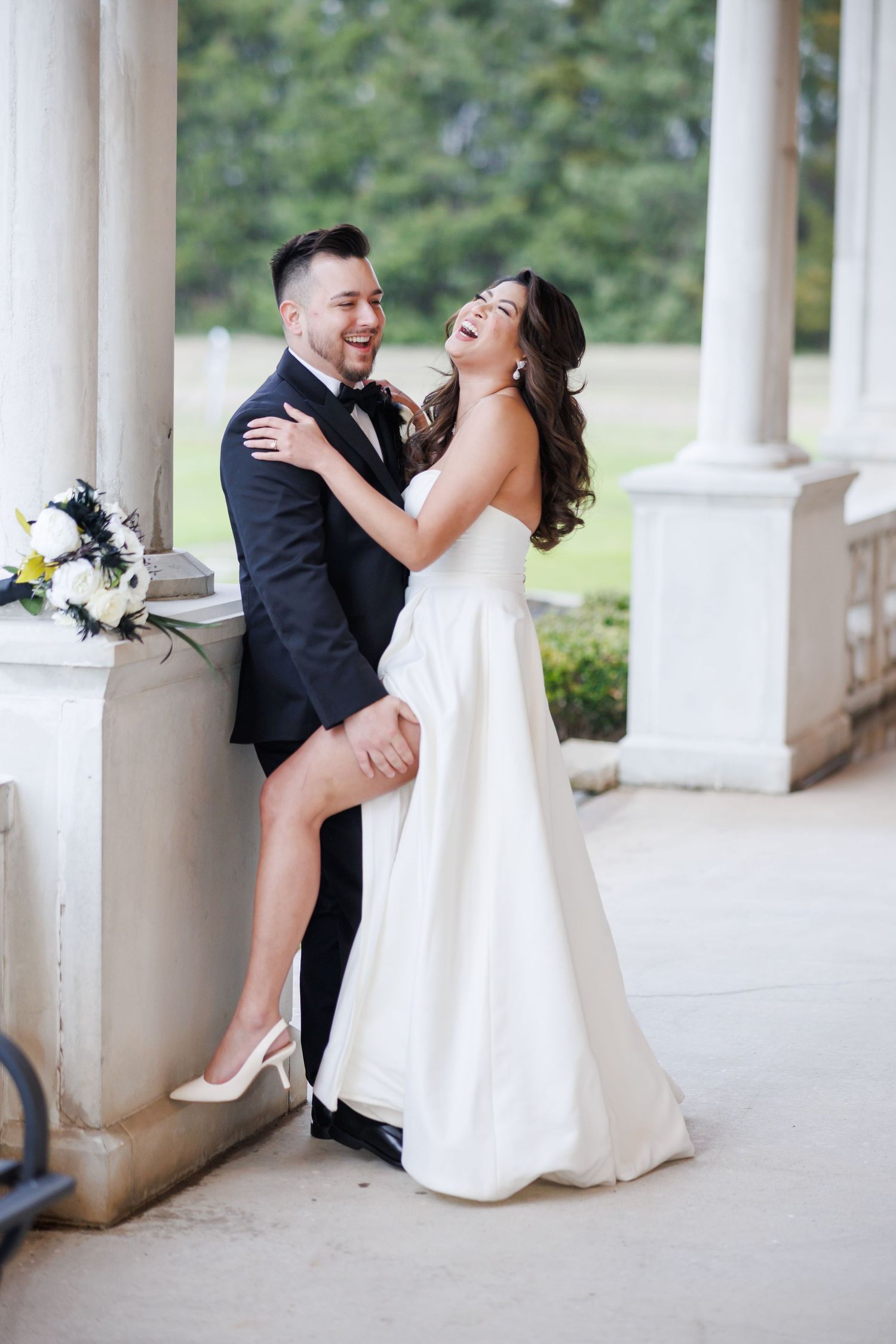 A joyful couple shares a candid moment laughing together during their wedding photoshoot near classic white columns.