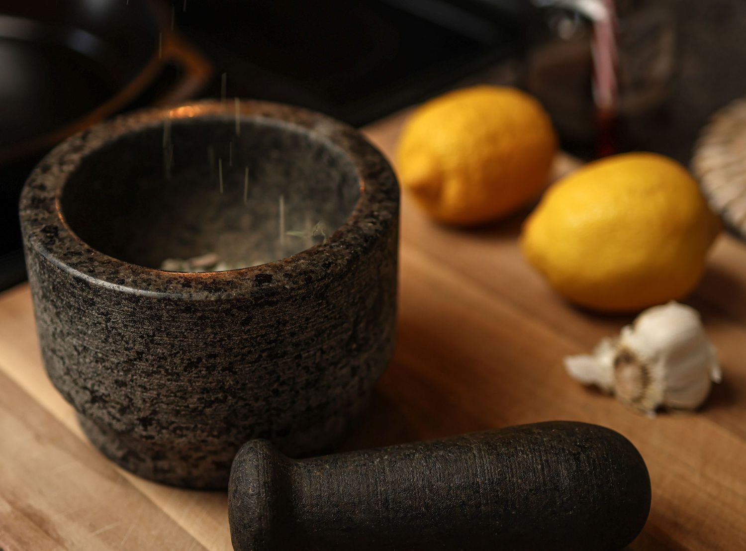 Dark stone mortar and pestle sits on wooden cutting board with fresh lemons and garlic cloves in background.