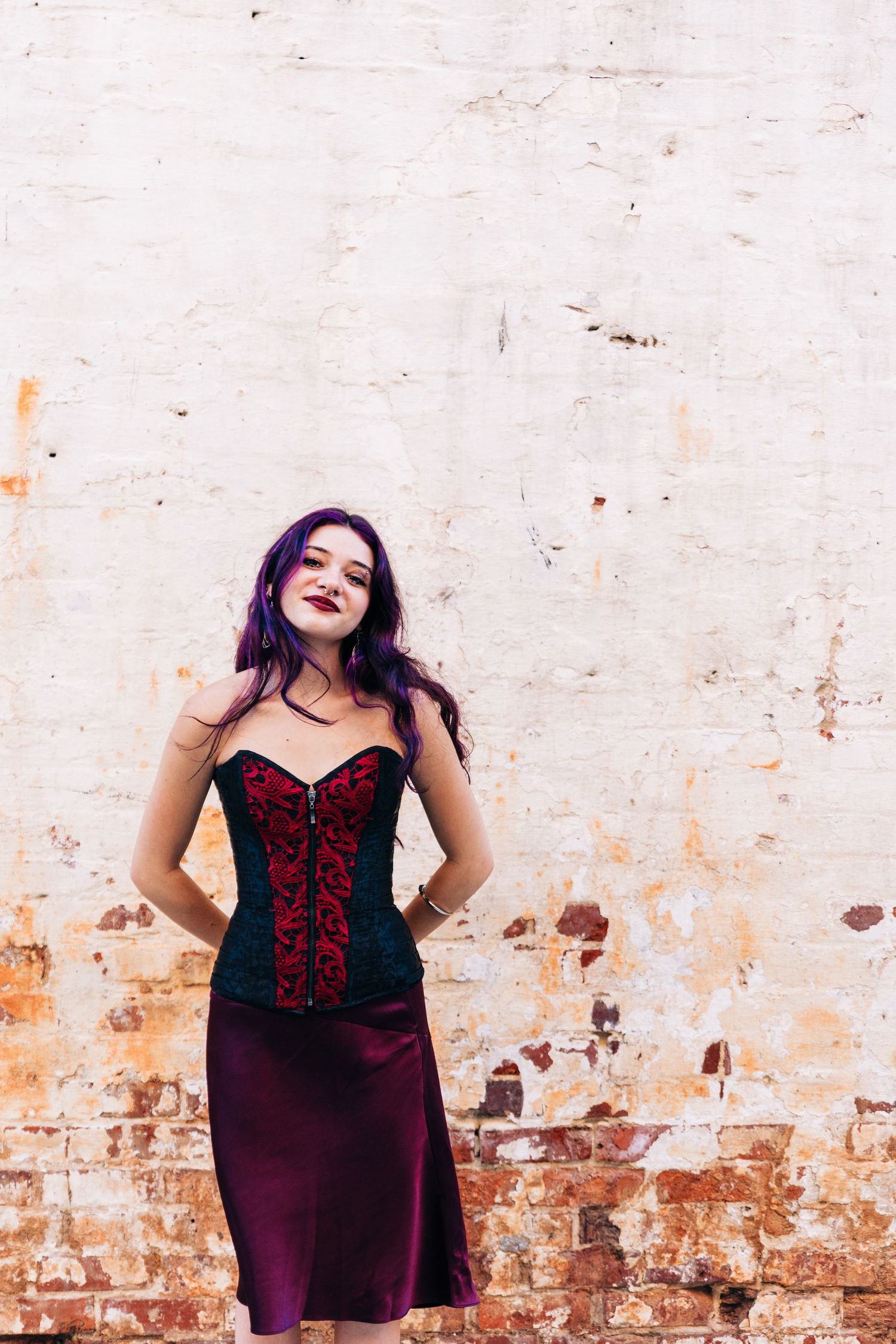 Gothic style outfit with red and black corset and burgundy skirt against weathered brick wall with faded white paint in Monroe, NC.
