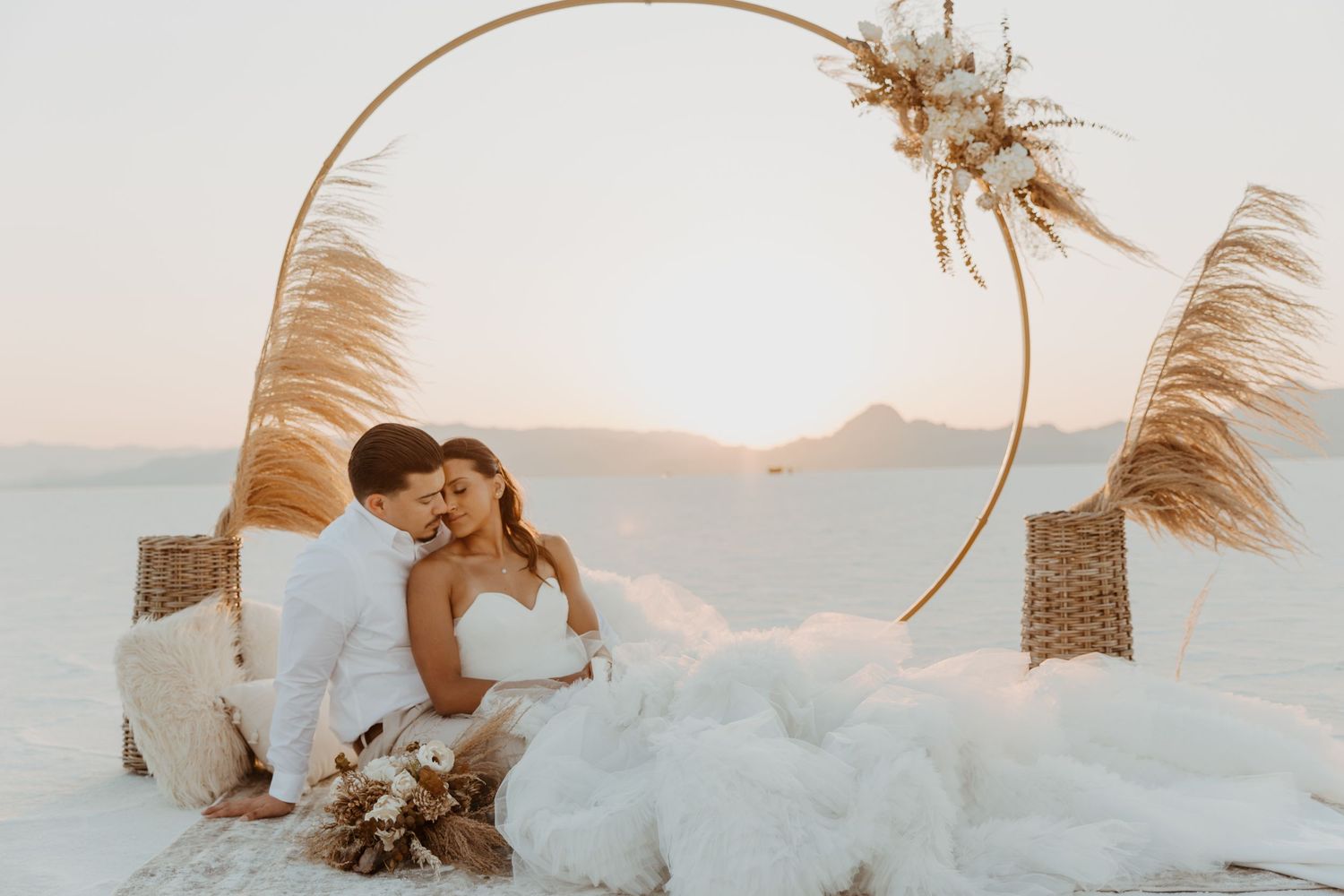 A romantic sunset beach scene with a circular arch decorated with pampas grass and dried flowers.