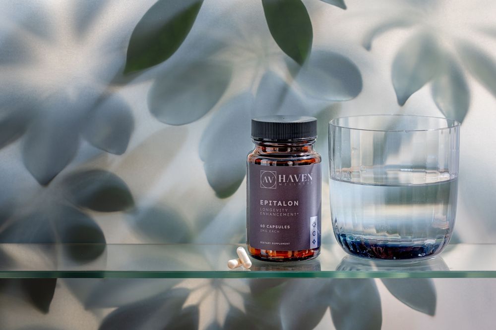 A bottle of red supplements sits next to a glass of water on a glass shelf with leaf shadows in the background.