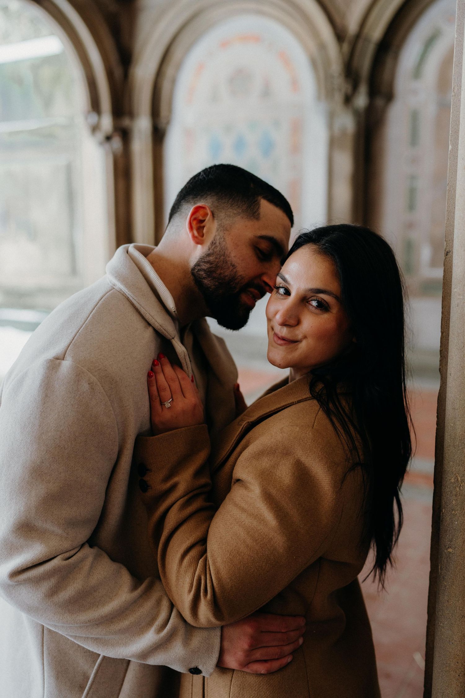 A romantic couple embracing under decorative arches in Bethesda Terrace in matching neutral-toned winter coats.