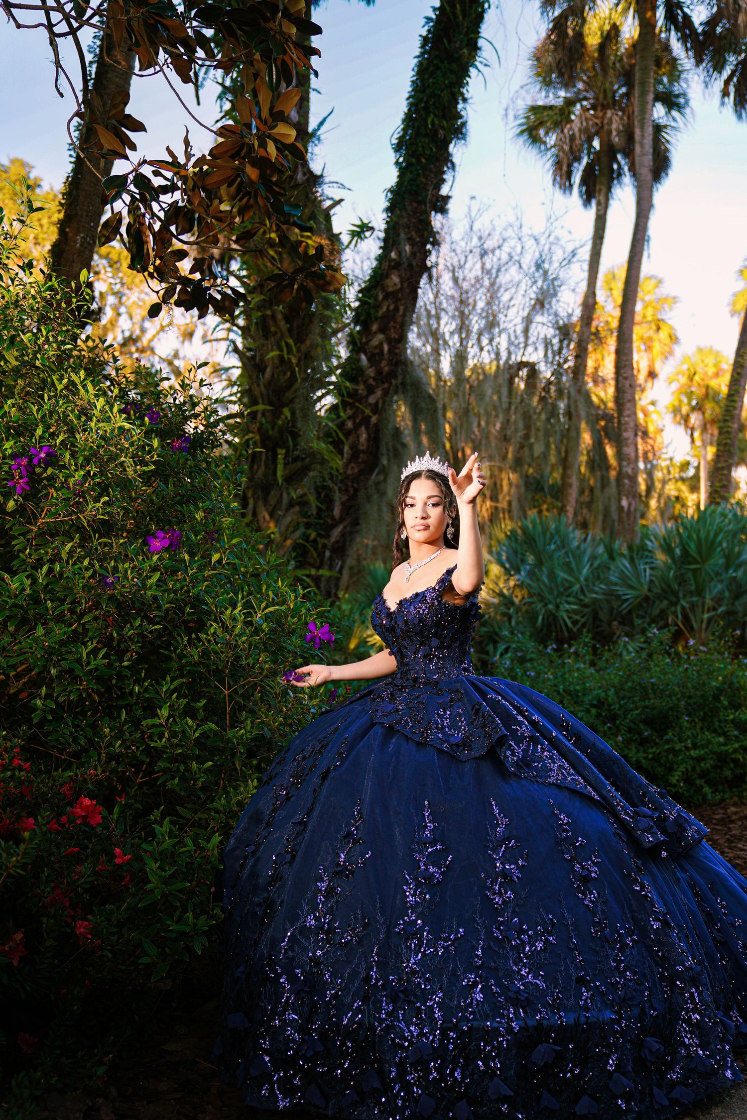 A person in a sparkling navy blue ball gown poses gracefully in a lush garden setting with palm trees at sunset.
