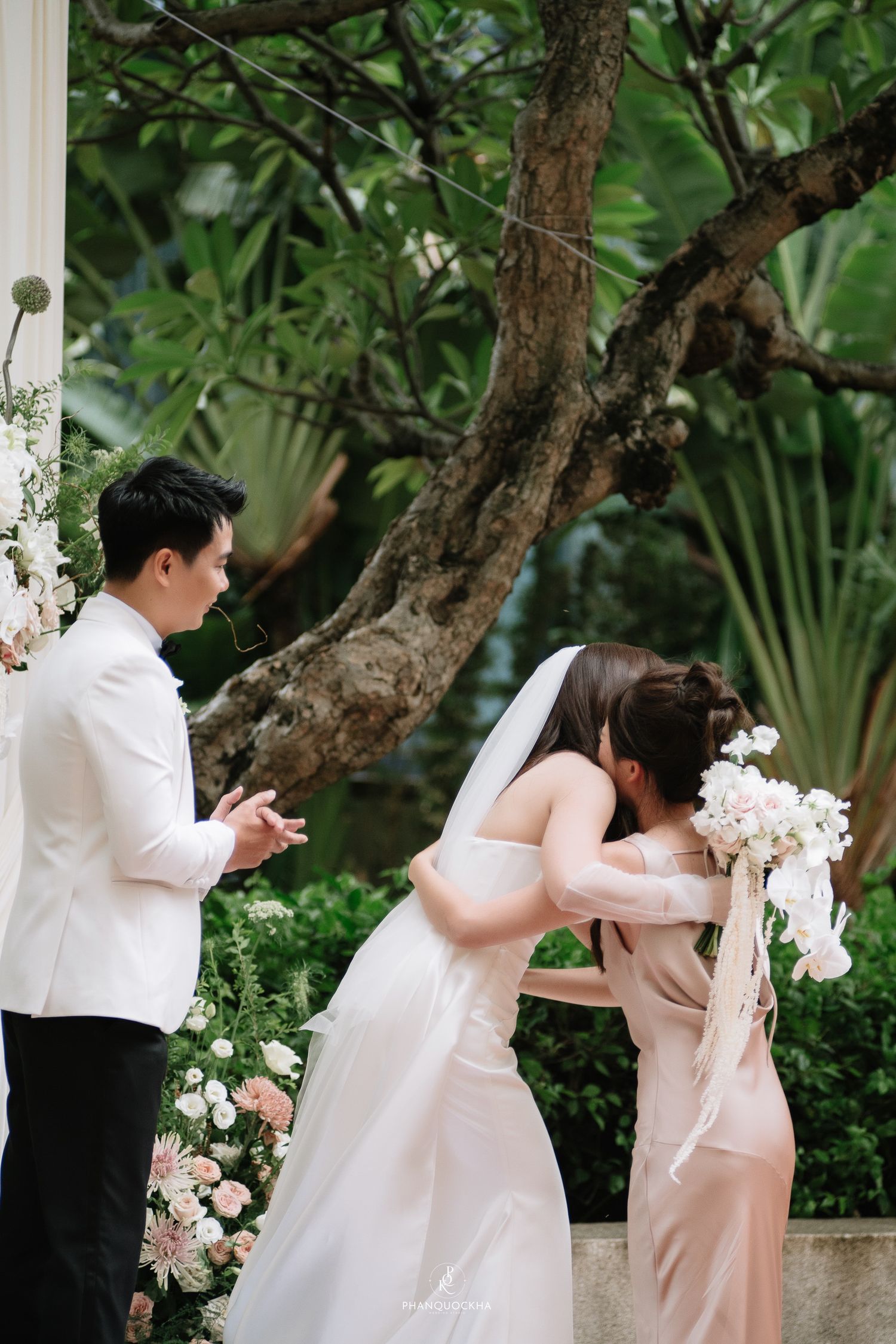 Bride in white gown embraces bridesmaid in blush dress while groom watches at outdoor garden wedding.